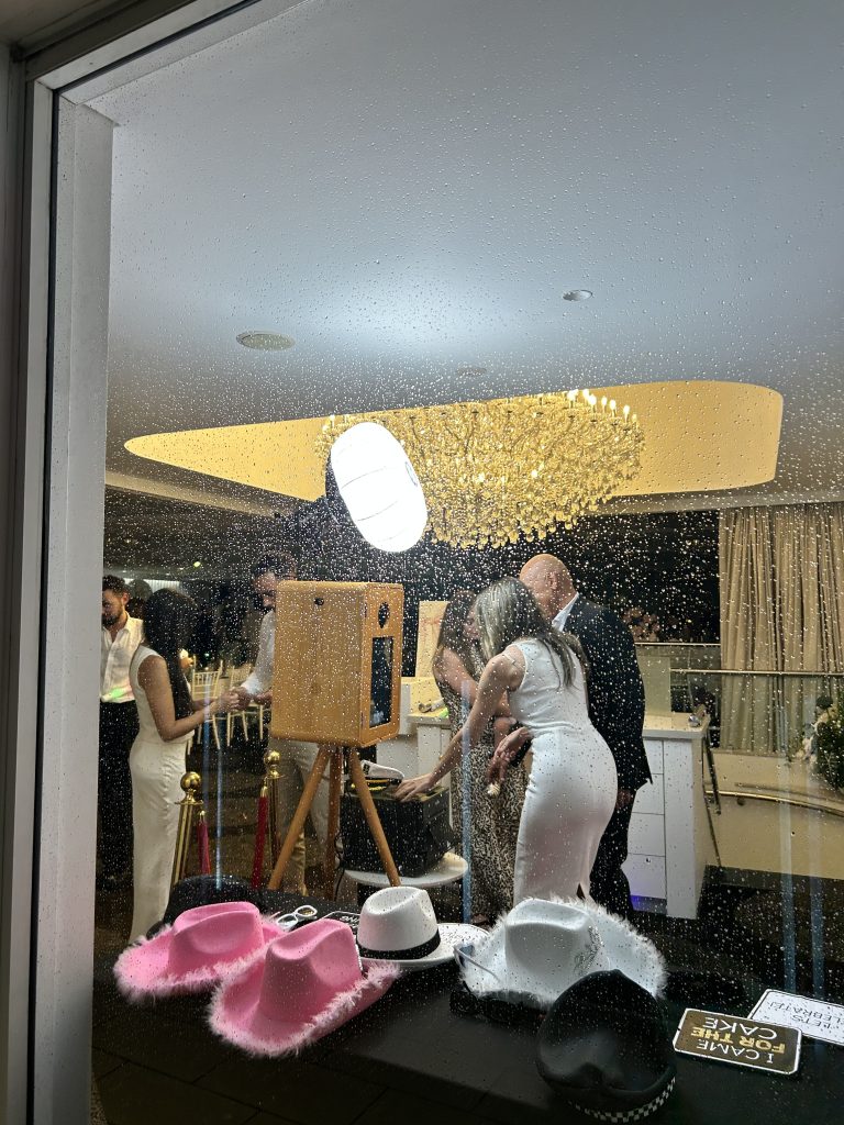 Party scene with wooden vintage photo booth setup and a couple that got their unlimited print copies and another couple operating the photobooth.