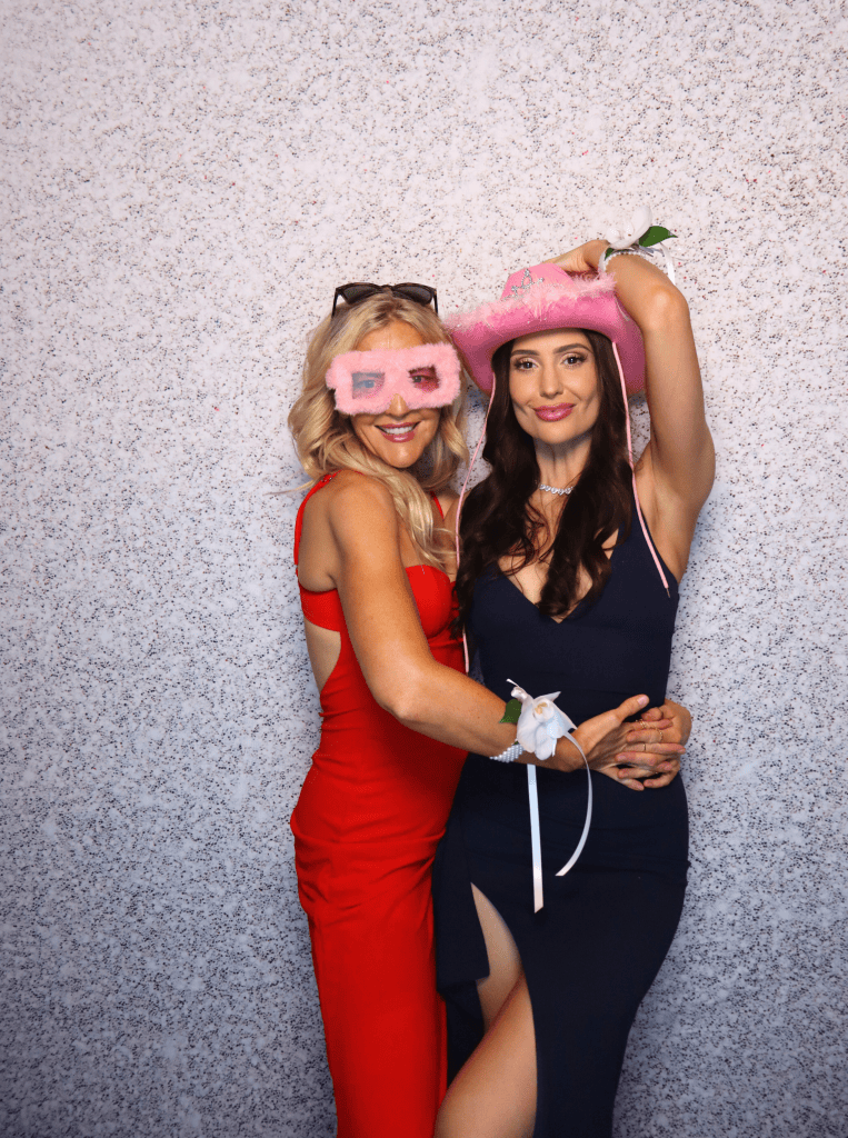Two young women posing for a photo with the mirror photobooth, wearing fun props in front of a silver backdrop.