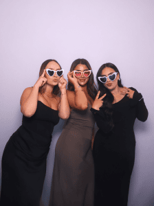 Group of 3 posing in front of the photo booth for a wedding all with glasses on.